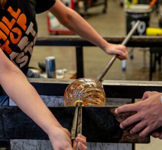 Close up image of a blown glass vessel being worked on by two Hilltop Artists students.