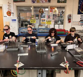 Photograph of four Hilltop Artists students working on the flameworking table in the hot shop at Hilltop Heritage Middle School