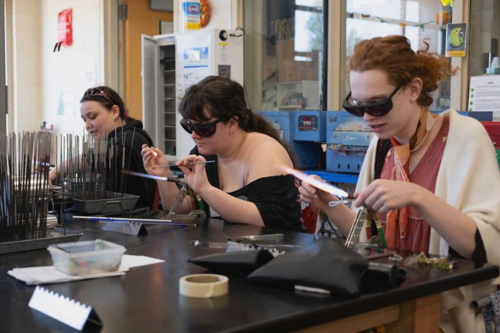 Group of three Hilltop Artists students working on the flameworking torches during a demonstration.